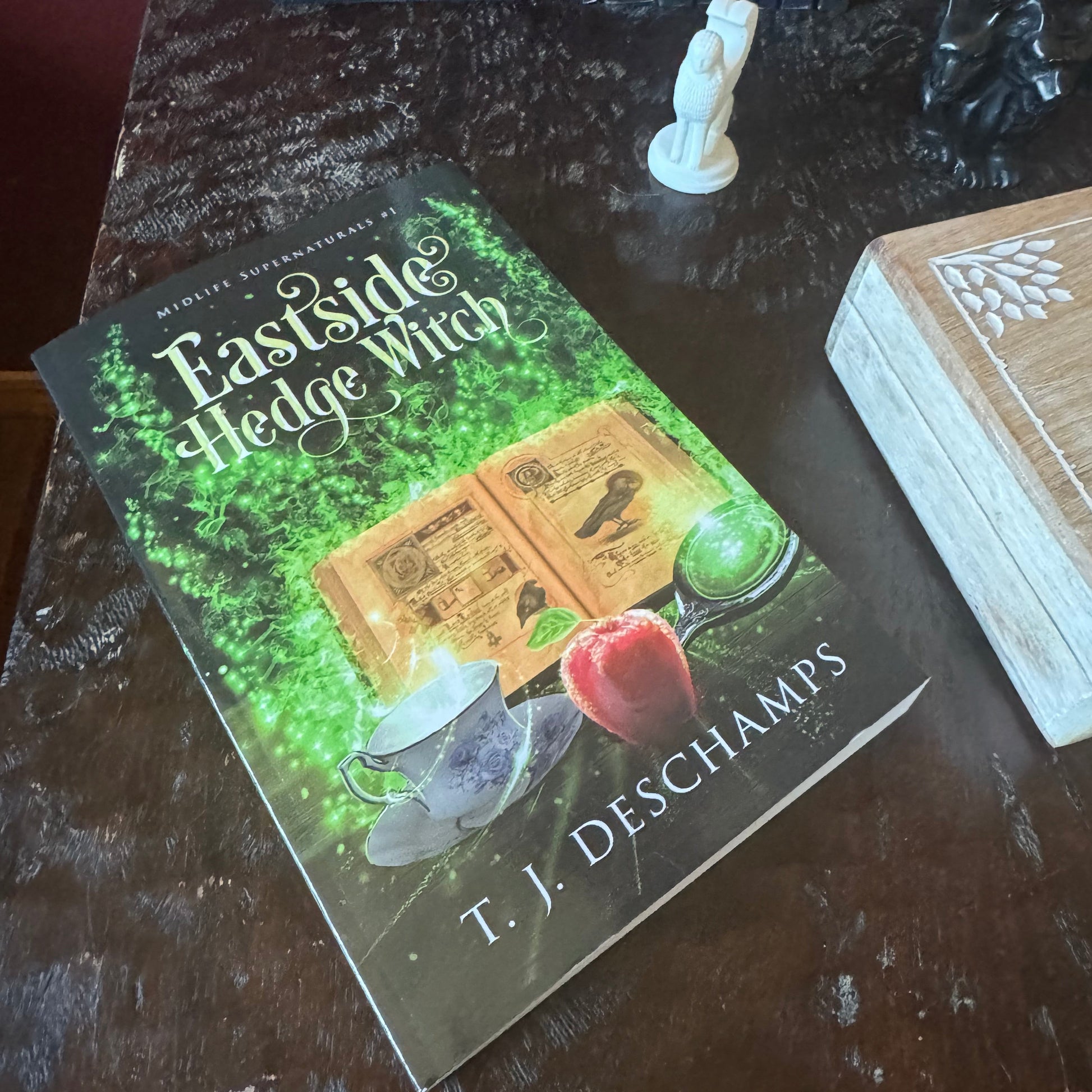 Sample paperback titled Eastside Hedge Witch by T.J. Deschamps with a picture of a grimoire, a teacup, an apple, and a magic mirror and magic flames. 